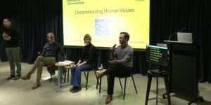 A photograph from the discussion panel. Three speakers sit on chairs next to a sign language interpreter and a screen showing an automatic transcription of the talk.