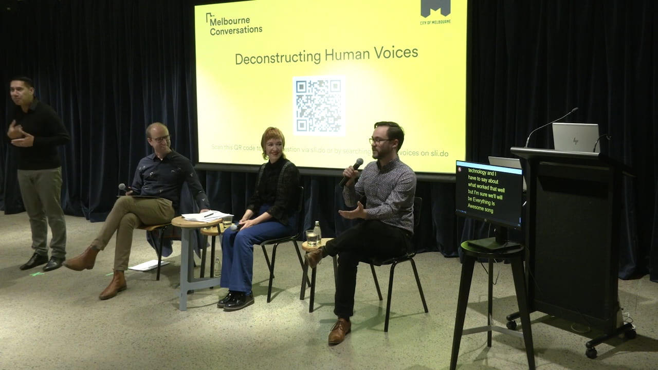 A photograph from the discussion panel. Three speakers sit on chairs next to a sign language interpreter and a screen showing an automatic transcription of the talk.